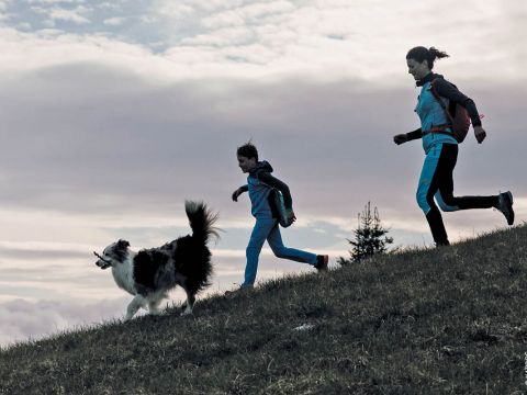 Eine Frau und ein Kind, beide in blauer Sportkleidung, laufen mit einem schwarz-weißen Hund, der einen Stock trägt, einen Grashang hinauf. Der Himmel ist mit weißen Wolken bedeckt. In der Ferne sind Bäume zu sehen.