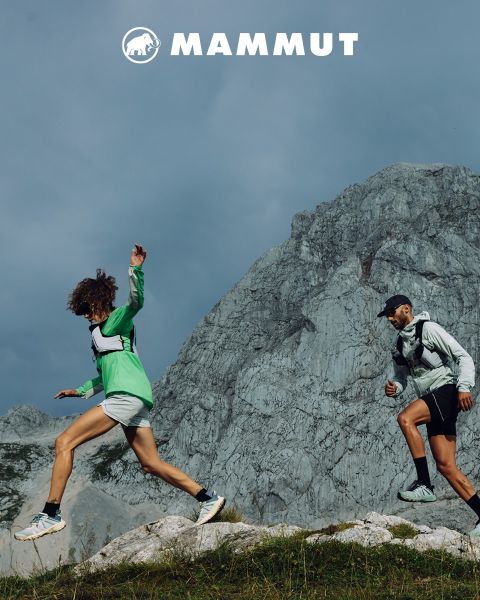 Zwei Wanderer in Outdoor-Ausrüstung durchqueren felsiges Gelände unter einem bewölkten Himmel, im Hintergrund ein großer Berg. Beide tragen Mammut-Kleidung, und das Markenlogo ist oben im Bild zu sehen.