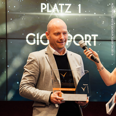 Eine Person im Anzug hält auf der Bühne eine Auszeichnung in die Höhe, neben ihr ein Mikrofon. Hinter ihr leuchtet ein Bildschirm mit den Aufschriften „PLATZ 1“ und „GIGASPORT“. Der dunkle Hintergrund mit hellen Punkten sorgt für eine festliche Atmosphäre.