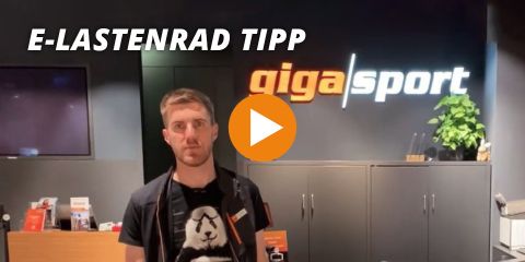 Eine Person steht in einem schwach beleuchteten Raum vor einer Wand mit dem Logo „gigasport“. Oben ist der Text „E-Lastenrad Tipp“ zu sehen. In der Bildmitte befindet sich ein großer orangefarbener Play-Button.