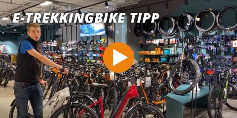Ein Fahrradladen mit einer Person neben einem E-Trekkingrad. An den Wänden des Ladens sind verschiedene Fahrräder, Zubehör und Reifen ausgestellt. Der Text „E-TREKKINGBIKE TIPP“ überlagert das Bild. In der Mitte befindet sich ein orangefarbener Play-Button.
