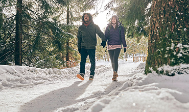 Ein Paar geht Hand in Hand durch einen verschneiten Waldweg, umgeben von hohen immergrünen Bäumen. Sie sind warm in Jacken, Mützen und Stiefel gekleidet und genießen einen sonnigen Wintertag.