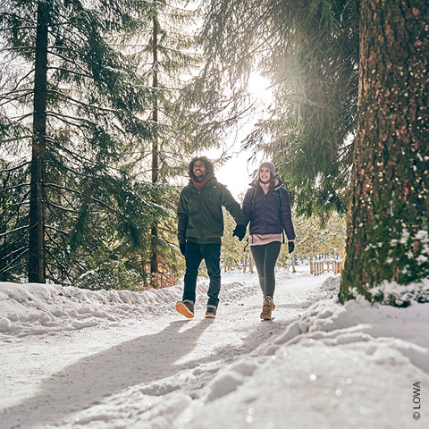 Ein Paar geht Händchen haltend einen schneebedeckten Waldweg entlang. Sonnenlicht fällt durch die hohen Bäume und schafft eine ruhige Winteratmosphäre. Beide tragen Wintermäntel und Stiefel und genießen gemeinsam die Natur.
