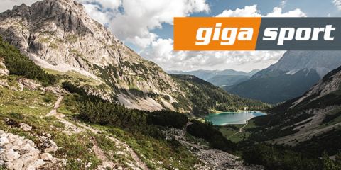 Malerische Berglandschaft mit felsigem Gipfel, viel Grün und einem kleinen See. Im Vordergrund schlängelt sich ein Wanderweg. Der Himmel ist teilweise bewölkt. Oben rechts befindet sich das „gigasport“-Logo.