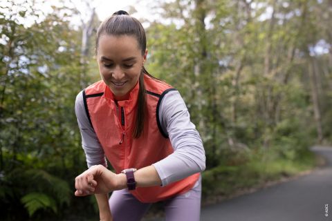 Eine Frau schaut beim Joggen im Freien auf ihre Smartwatch. Sie trägt ein rosa Unterhemd und ein graues Oberteil, umgeben von Grün und Bäumen. Ein Pfad schlängelt sich in die Ferne.