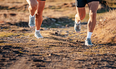 Zwei Personen laufen auf einem holprigen Pfad, nur ihre Beine sind zu sehen. Sie tragen Sportschuhe und Shorts. Der Boden ist uneben, mit Gras- und Schmutzflecken, was an eine natürliche Umgebung im Freien erinnert.