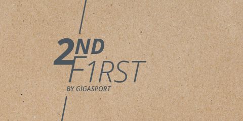 Das Bild zeigt die Wörter „2ND F1RST“ und „BY GIGASPORT“ in dunkler Schrift auf einem strukturierten, braunen, papierähnlichen Hintergrund. Eine dünne diagonale Linie trennt „2ND“ und „F1RST“.
