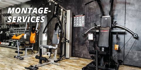 Ein Fitnessgeräte-Showroom mit verschiedenen Trainingsgeräten. Zu den Geräten gehören Hantelbänke, Kabelzugmaschinen und eine Kraftstation. In der oberen linken Ecke ist der Schriftzug „MONTAGE-SERVICES“ zu sehen. Der Boden ist aus Holz, die Wände dunkelgrau.