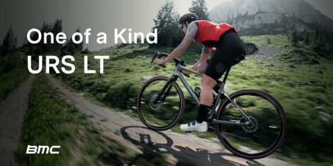 Ein Radfahrer in einem roten Oberteil fährt mit einem BMC URS LT einen grasbewachsenen Bergpfad hinauf. Im Hintergrund sind Berge und Bäume unter einem bewölkten Himmel zu sehen. Der Text auf dem Bild lautet: „Einzigartiges URS LT.“.