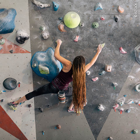 Eine Person mit langen Haaren klettert an einer Indoor-Boulderwand. Sie trägt ein Tanktop und hat einen Chalkbag an der Hüfte befestigt. Die Wand ist mit bunten Griffen in verschiedenen Formen und Größen ausgestattet.