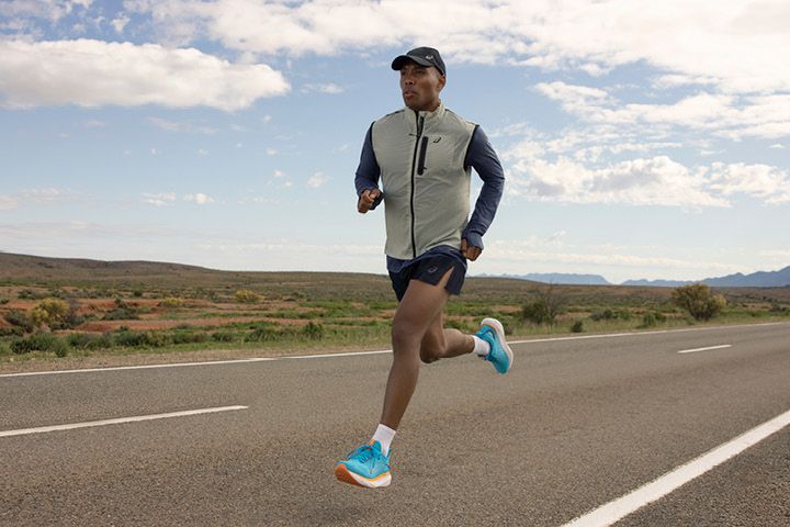 Eine Person joggt auf einer Landstraße. Sie trägt eine Mütze, eine leichte Weste und blaue Laufschuhe. Die Landschaft ist geprägt von offenen Feldern und Bergen in der Ferne unter einem teilweise bewölkten Himmel.