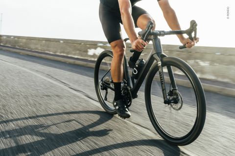 Eine Person in schwarzer Radsportkleidung fährt mit einem Rennrad schnell eine Straße mit niedriger Betonleitplanke entlang. Die Sonne wirft lange Schatten, was auf frühen Morgen oder späten Nachmittag hindeutet.