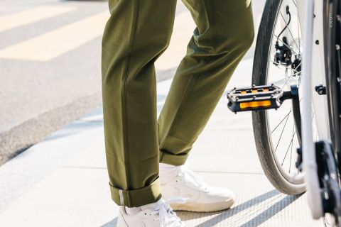 Eine Person in hochgekrempelten grünen Hosen und weißen Turnschuhen steht neben einem Fahrrad an einem Zebrastreifen. Der Fokus liegt auf den Unterschenkeln, den Schuhen und dem Fahrradpedal. Im Hintergrund ist die Straße zu sehen.