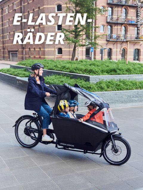 Eine Person mit Helm fährt ein schwarzes Lastenrad mit zwei Kindern im vorderen Bereich. Das Rad fährt auf einer gepflasterten Fläche in der Nähe eines Backsteingebäudes und einer Grünfläche. In der oberen linken Ecke ist der Schriftzug „E-Lastenräder“ zu sehen.