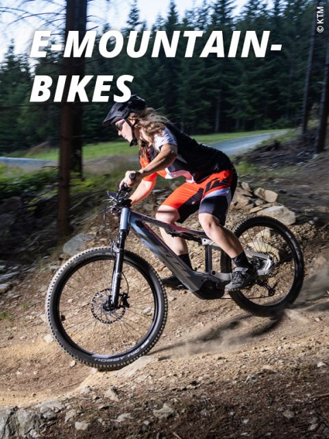 Eine Person fährt mit einem E-Mountainbike einen Waldweg hinunter. Sie trägt Helm, Sonnenbrille und rot-schwarzes Outfit. Sonnenlicht fällt durch die Bäume und beleuchtet den steinigen Weg. Die Beschriftung lautet „E-MOUNTAINBIKES“.