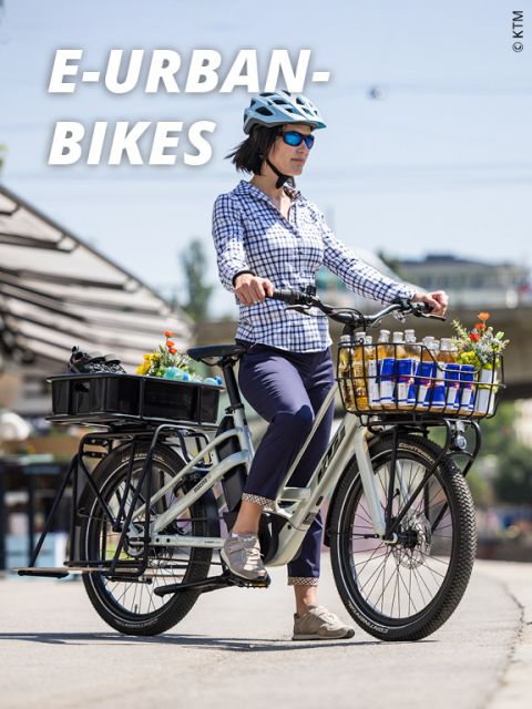 Eine Person mit Helm und Sonnenbrille fährt mit einem elektrischen Stadtfahrrad durch die Stadt. Das Fahrrad ist mit Körben gefüllt, die mit Getränkeflaschen und Blumen gefüllt sind. Der Schriftzug „E-URBAN-BIKES“ ist im Bild deutlich zu sehen.