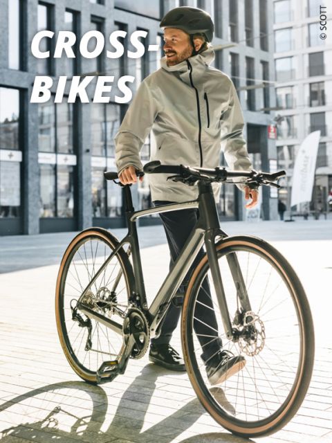 Eine Person mit Helm und leichter Jacke steht mit einem Fahrrad auf einer sonnenbeschienenen Straße. Im Hintergrund sind hohe, moderne Gebäude zu sehen. In der oberen linken Ecke steht der Text „CROSS-BIKES“.