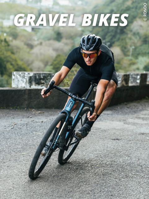 Radfahrer in schwarzer Kleidung und Helm lehnt sich auf einem Gravelbike in eine Kurve auf einer Schotterstraße. Im Hintergrund ist eine verschwommene, grüne Landschaft zu sehen. Oben steht der Text „GRAVEL BIKES“.