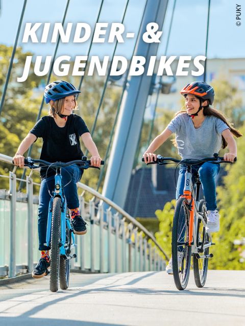 Zwei Kinder mit Helmen fahren mit dem Fahrrad über eine Brücke. Die Kinder lächeln und scheinen den sonnigen Tag zu genießen. Oben im Bild steht der Text „KINDER- & JUGENDBIKES“.