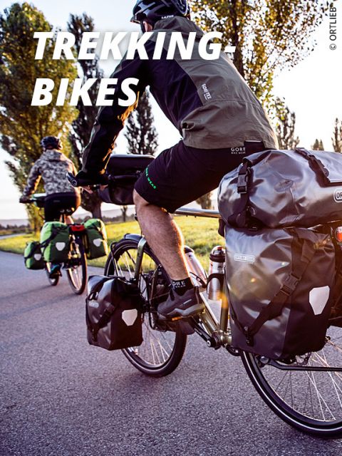 Zwei Radfahrer fahren mit beladenen Trekkingrädern und großen Packtaschen auf einer sonnigen Straße. Einer trägt eine schwarze Jacke, der andere eine Tarnjacke. Hohe Bäume säumen den Weg, und das Bild trägt die Aufschrift „Trekkingräder“.