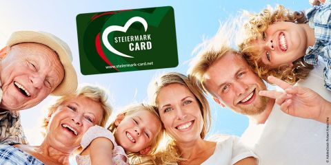 Eine Gruppe lächelnder Menschen, darunter Erwachsene und Kinder, steht im Kreis und blickt vor einem strahlend blauen Himmel in die Kamera. In den Himmel ist eine Grafik der Steiermark Card eingeblendet.