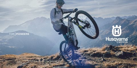 Ein Radfahrer macht auf einem Husqvarna E-Bike einen Wheelie auf einem Bergpfad. Er trägt Helm und Radsportbekleidung. Im Hintergrund sind schroffe Berge unter klarem Himmel zu sehen. Der Text lautet „Für Pioniere“ und zeigt das Husqvarna E-Bicycles-Logo.