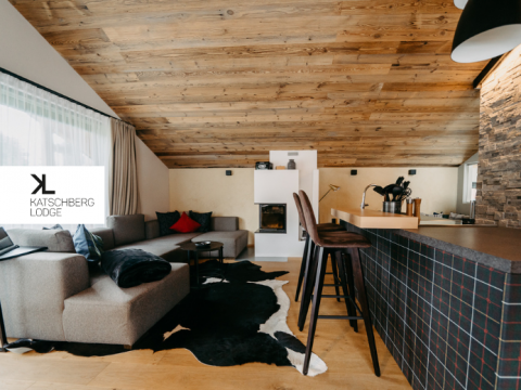 Ein gemütliches Wohnzimmer mit schräger Holzdecke. Es verfügt über ein graues Ecksofa, einen Kuhfellteppich und einen modernen Kamin. In der Nähe befindet sich eine Bar mit Hochstühlen. Die Wand ist teilweise mit Steinfliesen verkleidet. Ein Logo trägt die Aufschrift „Katschberg Lodge“.