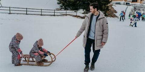 Ein Mann im Wintermantel zieht zwei Kinder auf einem Holzschlitten durch eine verschneite Landschaft. Die Kinder tragen gemusterte Schneeanzüge und rosa Mützen. Im Hintergrund fahren Menschen Ski.