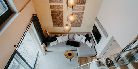 Ein gemütliches Wohnzimmer mit einem grauen L-förmigen Sofa und Kissen. Ein runder Couchtisch aus Holz steht auf einem hellen Teppich. Große Fenster mit Vorhängen sorgen für natürliches Licht. Hängelampen und abstrakte Wandkunst sorgen für Wärme und Stil.