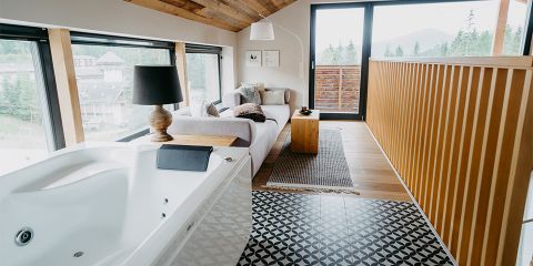 Ein gemütlicher Raum mit einem weißen Sofa, einer dekorativen Lampe und einem kleinen Holztisch auf einem gemusterten Teppich. Im Vordergrund steht ein großer Whirlpool. Der Raum besticht durch Holzakzente und große Fenster mit malerischem Blick auf Bäume und Hügel.