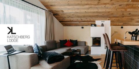 Gemütliches Interieur der Katschberg Lodge mit einem modernen, L-förmigen grauen Sofa, schwarzen und roten Kissen, einem weißen Kamin und einer hölzernen Frühstücksbar mit Hochstühlen. Der Raum hat beige Wände, eine Holzdecke und ein großes Fenster mit transparenten Vorhängen.