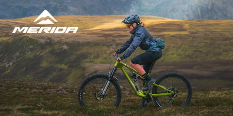 Ein Radfahrer fährt mit seinem grünen Mountainbike einen grasbewachsenen Hang entlang. Er trägt Helm und Outdoor-Ausrüstung. Die Landschaft ist von sanften Hügeln und einem teilweise bewölkten Himmel geprägt. In der oberen linken Ecke erscheint das Wort „Merida“ mit einem Berglogo.