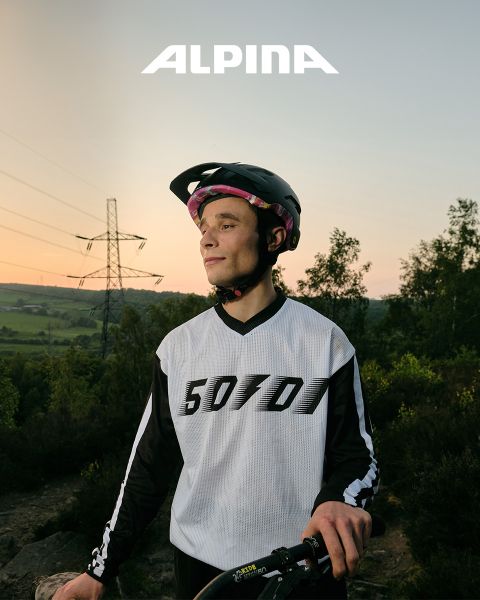 Ein Radfahrer mit Helm und weißem Hemd steht mit seinem Fahrrad da und blickt bei Sonnenuntergang in die Ferne. Im Hintergrund sind Bäume, Hügel und eine Stromleitung zu sehen. Darüber ist das Wort „ALPINA“ zu sehen.