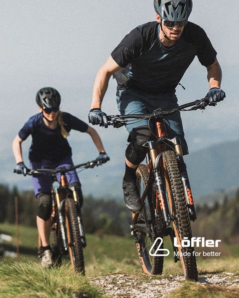 Zwei Mountainbiker fahren auf einem felsigen Trail inmitten üppiger Vegetation. Beide tragen Helme und Sonnenbrillen. In der unteren rechten Ecke ist der Schriftzug „Löffler Made for better“ zu sehen.