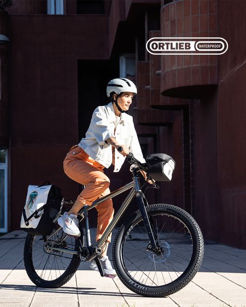 Eine Person mit Helm und Freizeitkleidung fährt mit einem Fahrrad, an dem wasserdichte Ortlieb-Taschen befestigt sind, in der Nähe moderner Backsteingebäude. Die Sonne wirft lange Schatten und unterstreicht ihren konzentrierten Gesichtsausdruck.