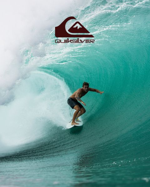 Ein Surfer reitet auf einer großen türkisfarbenen Welle und verkörpert Balance und Können. Er trägt dunkle Badeshorts, und die Welle kräuselt sich über ihm. Über ihm ist das Quiksilver-Logo zu sehen.
