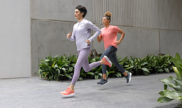 Zwei Frauen joggen nebeneinander in einer städtischen Umgebung. Die eine trägt ein hellviolettes Outfit mit rosa Schuhen, die andere ein rosa Oberteil mit dunklen Leggings. Grüne Pflanzen säumen den Weg, im Hintergrund ist eine Betonmauer zu sehen.