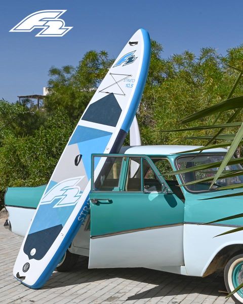 Ein türkis-weißer Oldtimer steht geparkt, an dem ein blau-weißes Paddleboard lehnt. Im Hintergrund sind grüne Bäume zu sehen, und in der oberen linken Ecke befindet sich ein Logo.