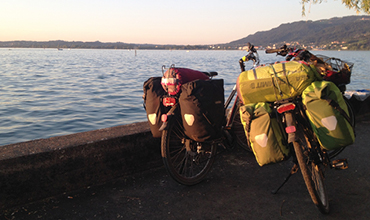 Zwei Fahrräder mit beladenen Packtaschen stehen an einem Uferweg. Die untergehende Sonne taucht die Szene in ein warmes Licht. Im Hintergrund sind ruhiges Wasser und ferne Hügel zu sehen.