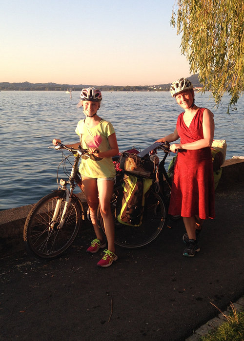 Zwei Personen mit Helmen stehen bei Sonnenuntergang neben ihren Fahrrädern auf einem Seeuferweg. Die eine trägt ein rotes Kleid, die andere ein helles Outfit. Beide haben Fahrradtaschen an ihren Rädern. Im Hintergrund sind der See und die Bäume zu sehen.