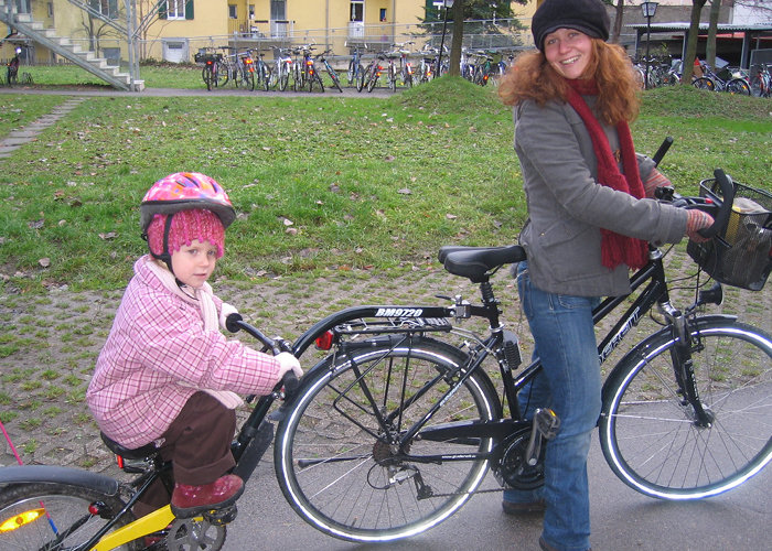 Eine Frau und ein kleines Kind stehen neben ihren Fahrrädern. Das Kind sitzt auf einem kleineren Fahrrad, das an das größere angeschlossen ist. Beide sind warm angezogen, das Kind trägt einen Helm. Im Hintergrund sind abgestellte Fahrräder und Gebäude zu sehen.
