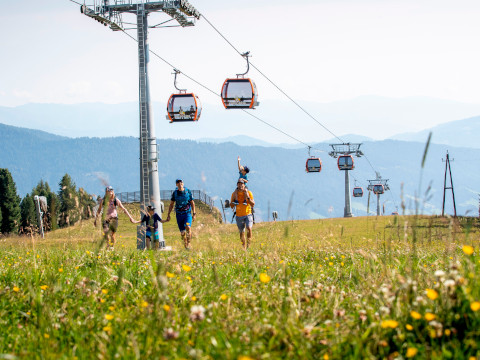 Eine vierköpfige Familie geht Hand in Hand über eine Wiese mit Wildblumen, unter einem Skilift mit orangefarbenen Gondeln hindurch. Im Hintergrund sind Berge unter einem klaren Himmel zu sehen.