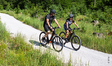 Zwei Radfahrer, mit Helmen und Sportkleidung, fahren auf einem Schotterweg, umgeben von üppigem Grün und Bäumen. Der Weg schlängelt sich durch ein Waldgebiet, und beide Radfahrer scheinen den sonnigen Tag und die malerische Umgebung zu genießen.