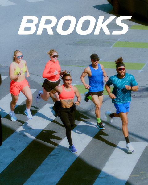 Eine Gruppe von fünf Personen rennt energisch über einen Zebrastreifen. Sie tragen farbenfrohe Sportkleidung, darunter Shorts und Tanktops. Die Szene ist lebhaft, über ihnen prangt deutlich das Wort „BROOKS“.