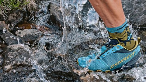 Der Fuß eines Läufers watet durch einen Bach. Er trägt blaue Dynafit-Trailschuhe und bunte Socken. Wassertropfen und Steine sind sichtbar und unterstreichen die Bewegung und das raue Gelände.