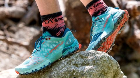 Nahaufnahme einer Person in blaugrünen Trailrunning-Schuhen mit filigranem Profil, die über einen Felsen steigt. Die Person trägt außerdem rosa und schwarze Socken. Ein Teil des natürlichen Trail-Hintergrunds ist sichtbar.