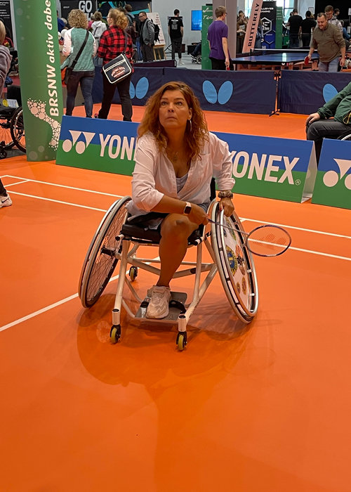 Eine Frau im Rollstuhl steht mit einem Schläger in der Hand auf einem orangefarbenen Badmintonfeld. Sie scheint sich auf das Spiel zu konzentrieren. Menschen haben sich um das Feld versammelt, im Hintergrund sind Sportbanner zu sehen.