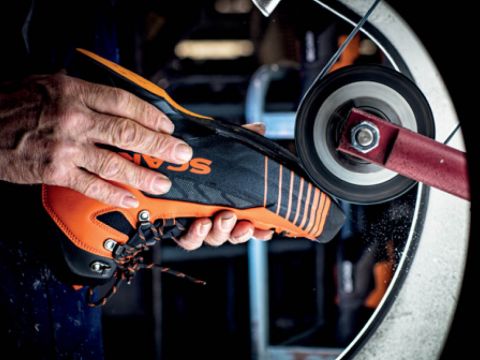 Eine Person hält einen orange-schwarzen Schuh an eine Schleifscheibe und glättet oder poliert ihn. Der Schuh hat Schnürsenkel und wird in einer Werkstatt oder Fabrik bearbeitet.