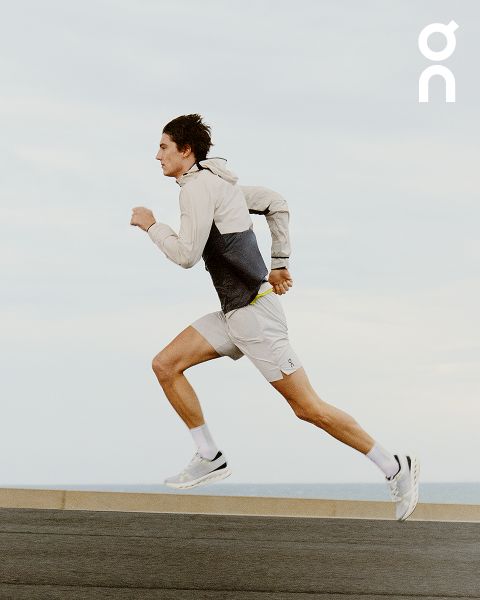 Eine Person läuft im Freien. Sie trägt eine beige-graue Windjacke, hellgraue Shorts und weiße Laufschuhe. Der Hintergrund ist ein klarer Himmel und eine ebene Fläche. Das Logo „On“ ist in der oberen rechten Ecke sichtbar.