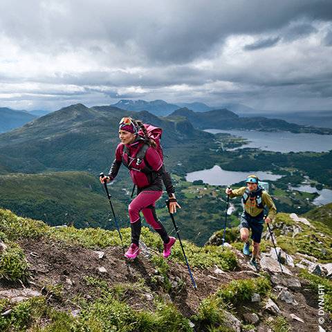 Zwei Wanderer mit Rucksäcken und Trekkingstöcken steigen einen grasbewachsenen Bergpfad hinauf. Der Wanderer im Vordergrund trägt Rosa und Lila, der dahinter Grün und Blau. Unter einem dramatischen Wolkenhimmel bietet sich ein malerischer Blick auf Täler, Seen und ferne Berge.